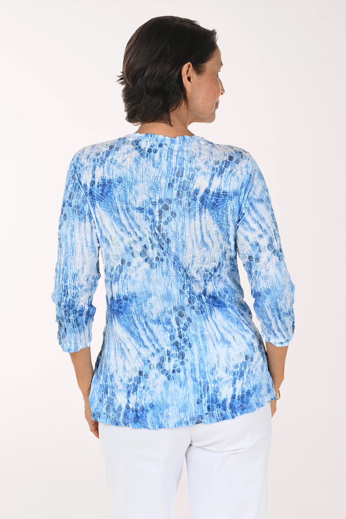 A smiling woman wearing a blue and white tie-dye patterned wrap top with three-quarter sleeves and white pants, photographed against a plain light background.