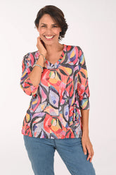A smiling woman wearing a colorful abstract print top with three-quarter sleeves and blue jeans, standing against a plain white studio background.