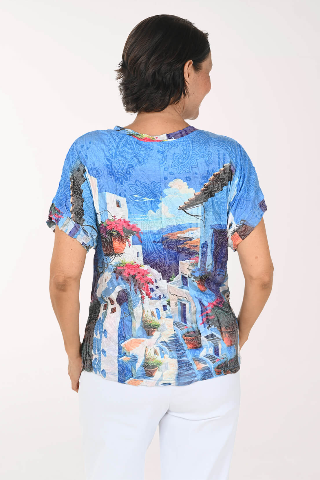 A woman viewed from the back wearing a short-sleeve crewneck top with a vibrant blue and white Mediterranean village print, paired with white pants, against a plain white background.