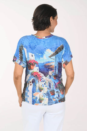 A woman viewed from the back wearing a short-sleeve crewneck top with a vibrant blue and white Mediterranean village print, paired with white pants, against a plain white background.