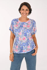A smiling woman wearing a short-sleeve blue and pink floral print top with blue jeans, standing against a plain white studio background.