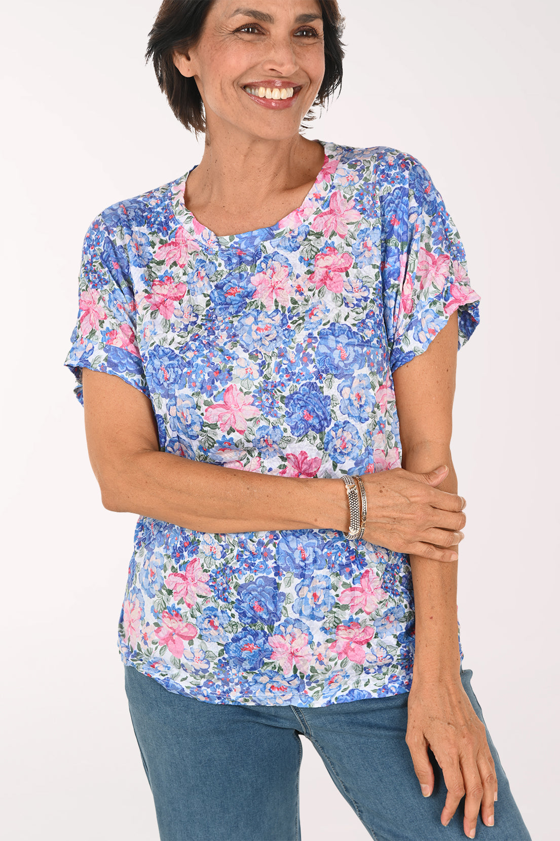 A smiling woman wearing a short-sleeve blue and pink floral print top with blue jeans, standing against a plain white studio background.