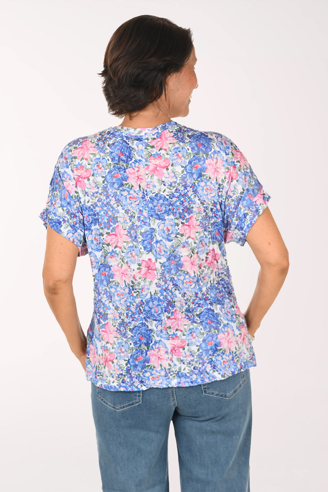A smiling woman wearing a short-sleeve blue and pink floral print top with blue jeans, standing against a plain white studio background.
