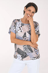 A smiling woman wearing a short-sleeve blouse with a multicolor abstract patchwork print in shades of blue, gray, black, and beige, paired with white pants, against a plain white studio background.