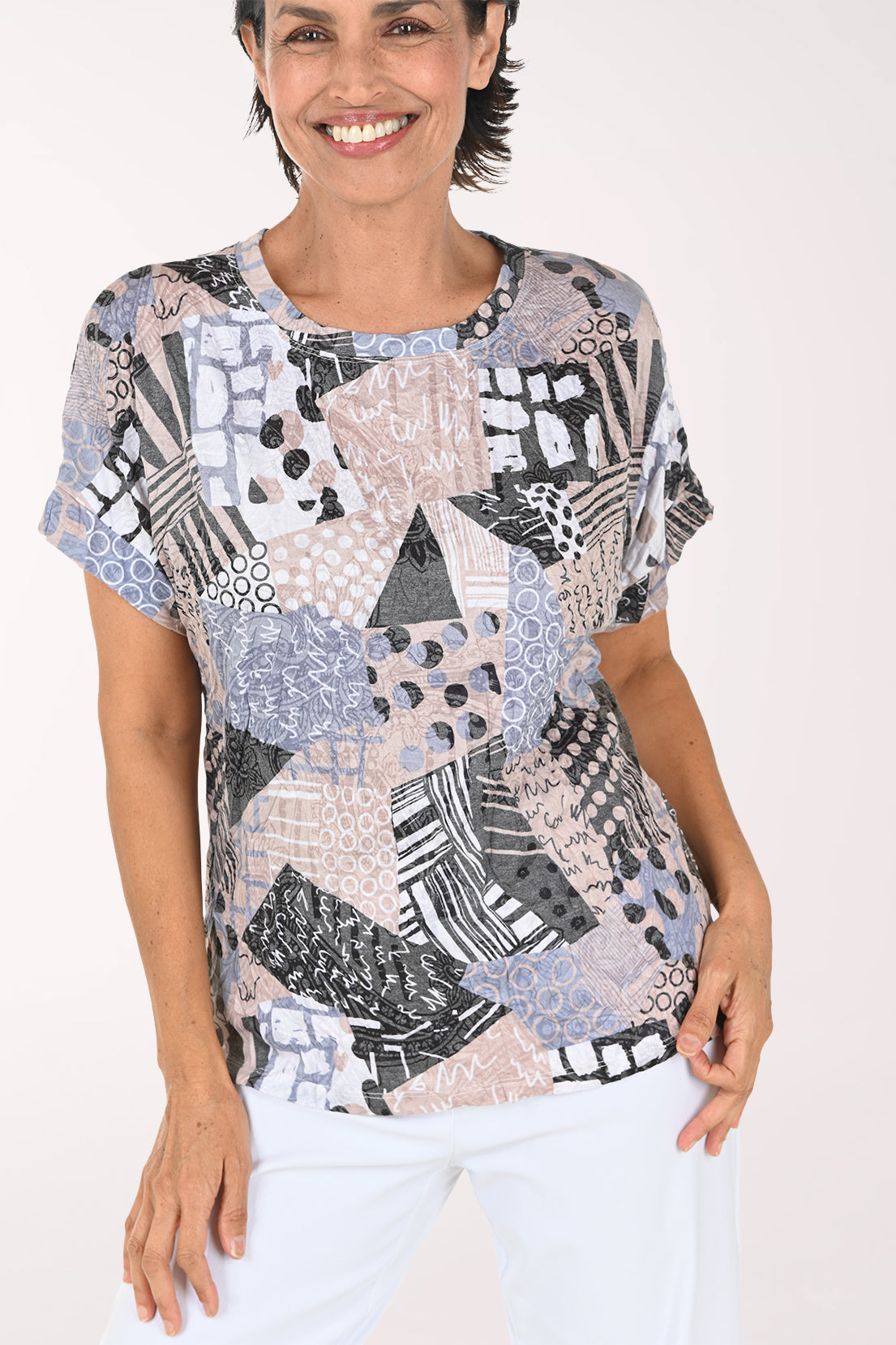 A smiling woman wearing a short-sleeve blouse with a multicolor abstract patchwork print in shades of blue, gray, black, and beige, paired with white pants, against a plain white studio background.