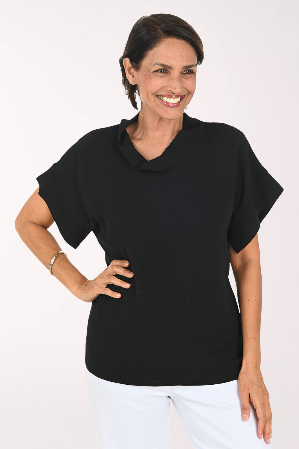 A smiling woman wearing a black short-sleeve cowl-neck top and white pants, standing against a plain white studio background.