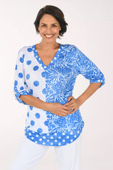 A woman modeling a blue and white patterned top with three-quarter sleeves and a V-neck, paired with white pants, against a plain white studio background.