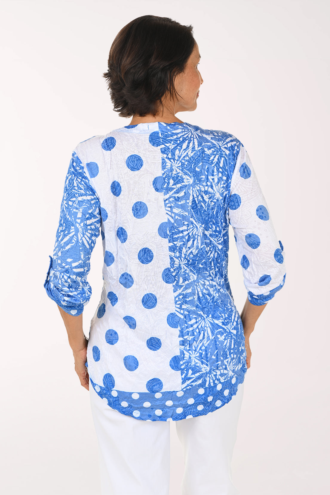 A woman modeling a blue and white patterned top with three-quarter sleeves and a V-neck, paired with white pants, against a plain white studio background.