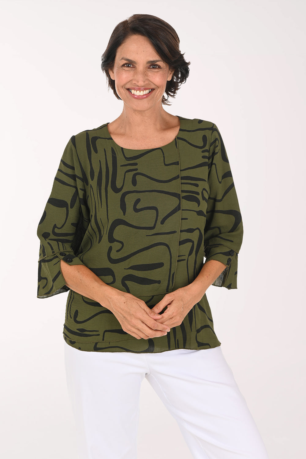 A smiling woman wearing an olive green and black abstract patterned blouse with three-quarter ruffled sleeves, paired with white pants, against a plain white studio background.
