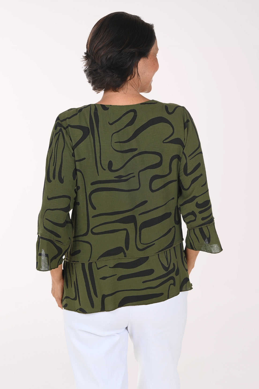 A smiling woman wearing an olive green and black abstract patterned blouse with three-quarter ruffled sleeves, paired with white pants, against a plain white studio background.