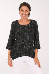 A woman wearing a black three-quarter sleeve top with a white geometric line pattern, paired with white pants, standing against a plain white background.