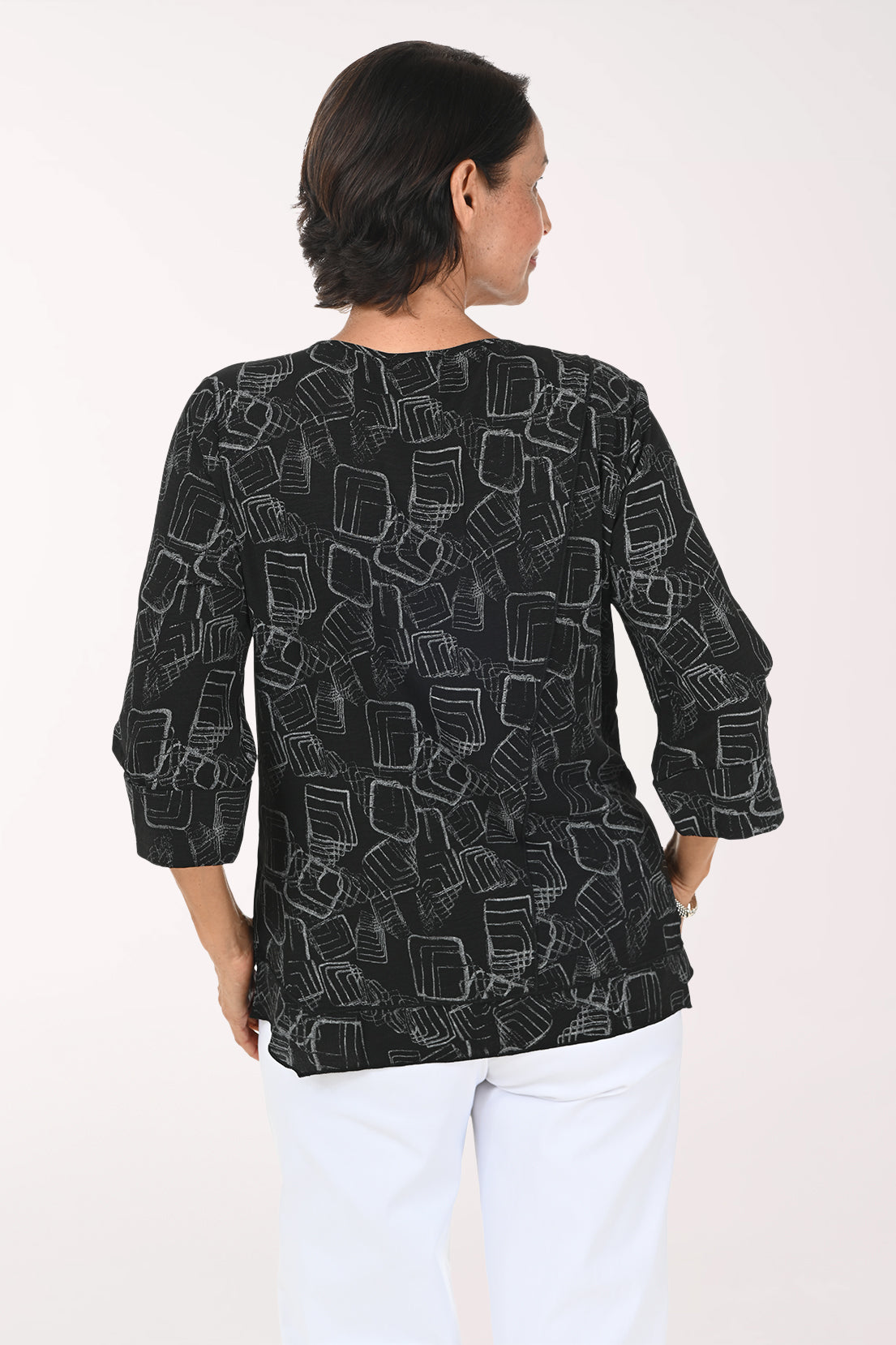 A woman wearing a black three-quarter sleeve top with a white geometric line pattern, paired with white pants, standing against a plain white background.