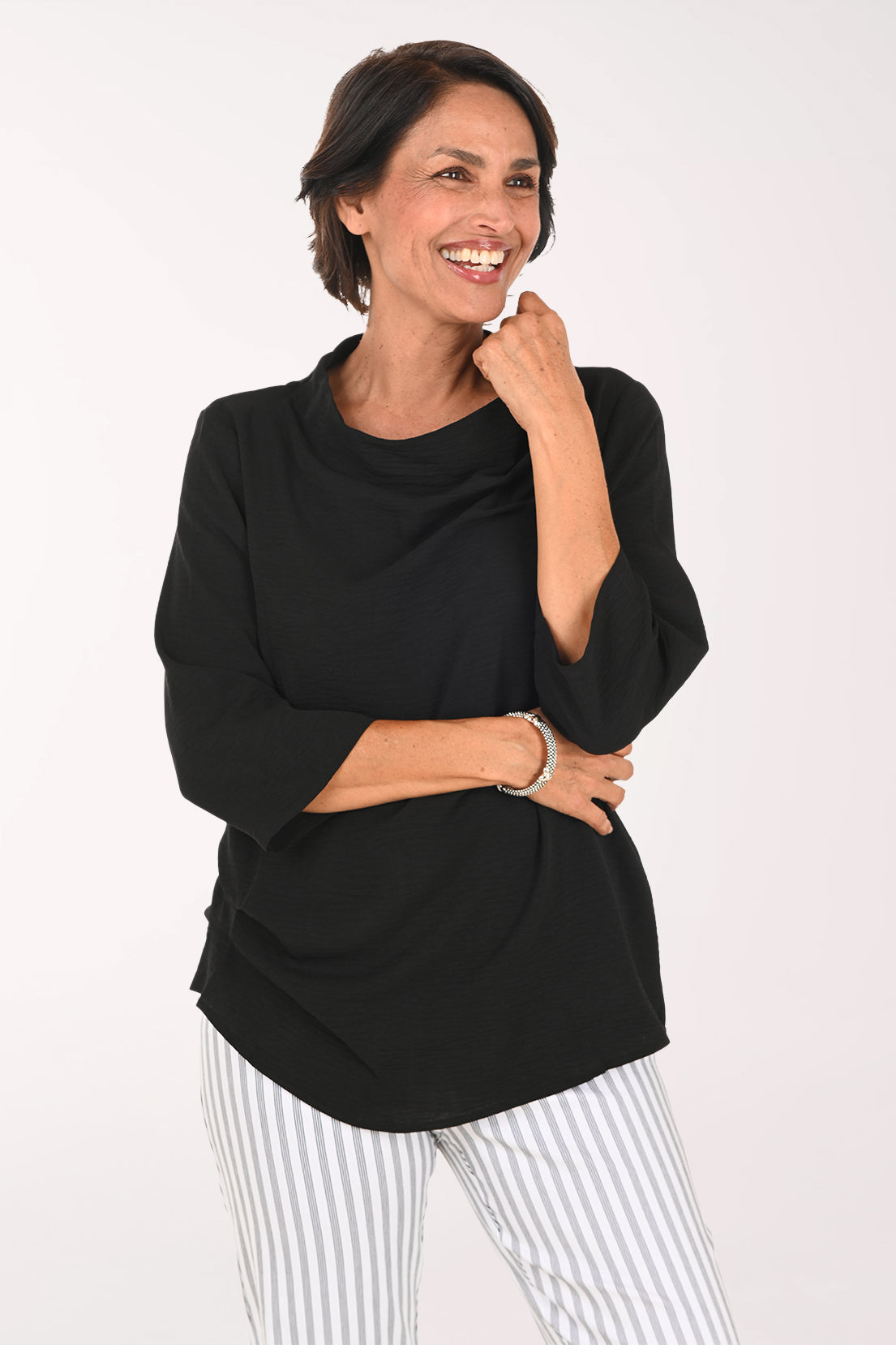 A smiling woman with short brown hair wearing a black cowl-neck three-quarter sleeve top and white and gray vertically striped pants, posing against a plain white studio background.