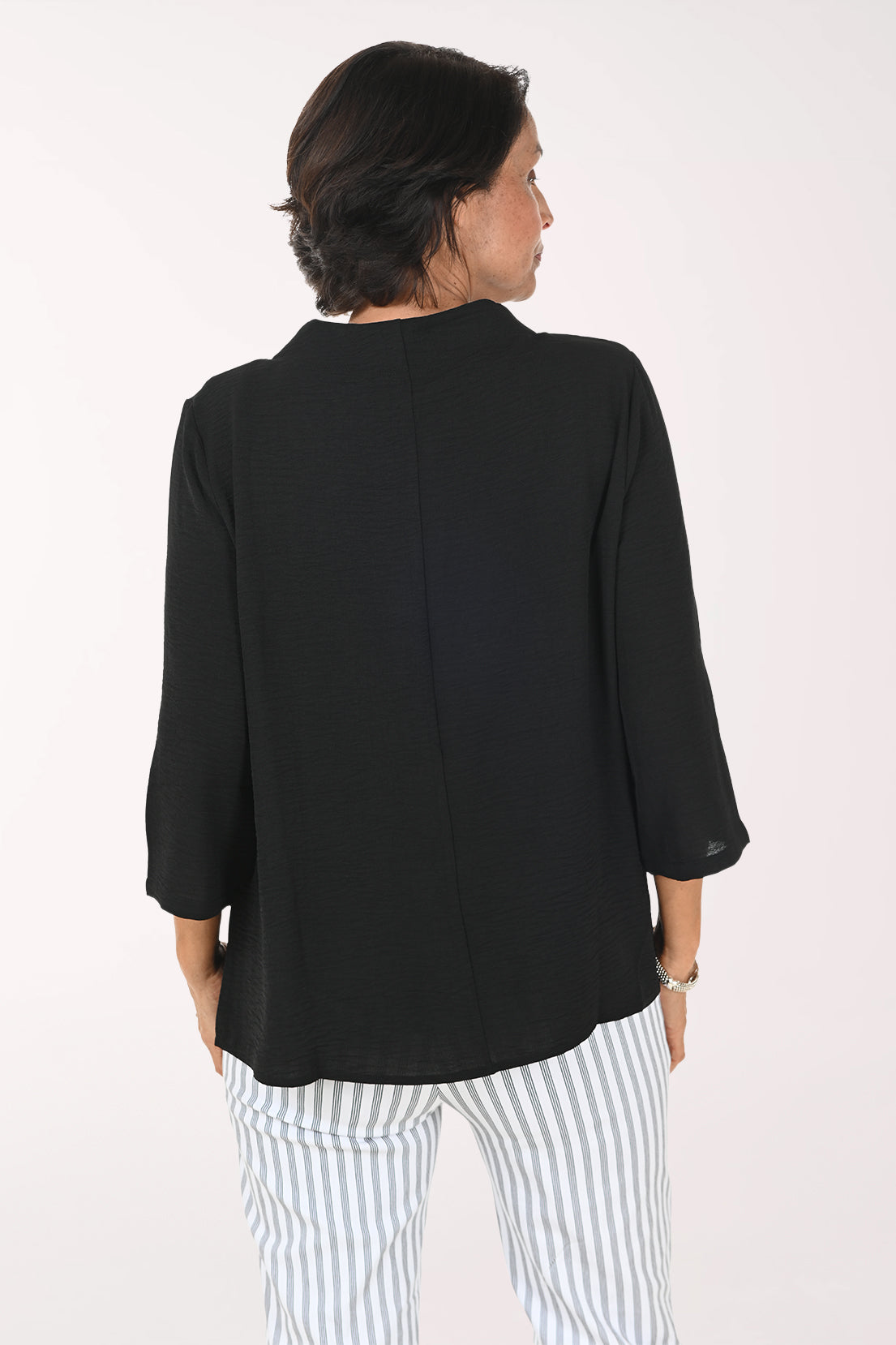 A smiling woman with short brown hair wearing a black cowl-neck three-quarter sleeve top and white and gray vertically striped pants, posing against a plain white studio background.