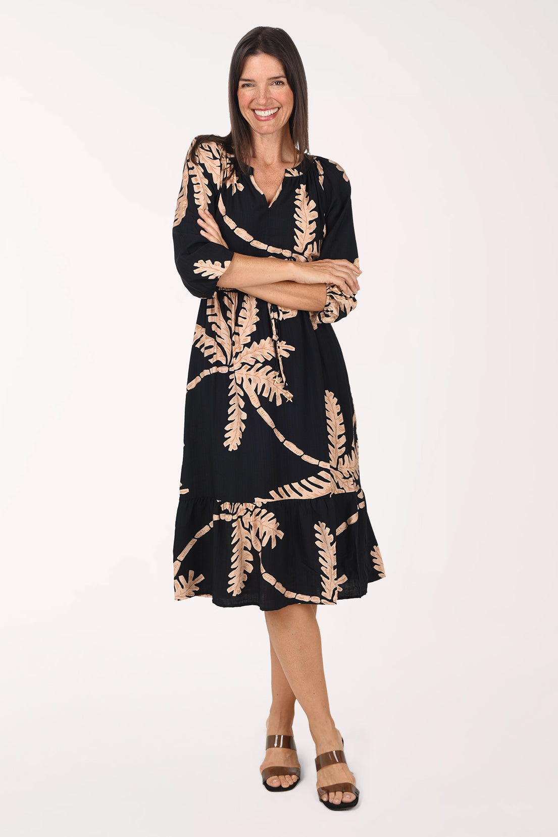 Model wearing the Mlle Gabrielle Coconut Palm Cinched Waist Midi Dress, a black midi dress with a taupe palm tree print, front view