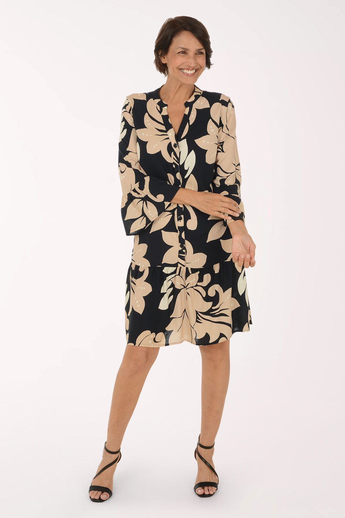 A woman modeling a knee-length black and beige floral print dress with three-quarter bell sleeves and a button-up front, paired with black strappy high-heeled sandals, against a plain white studio background.
