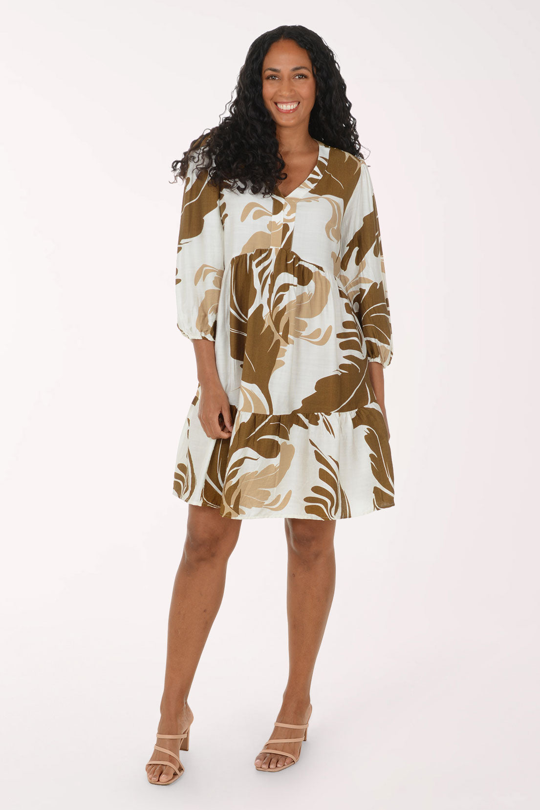 A woman modeling a short tiered dress with three-quarter sleeves, featuring an abstract botanical pattern in brown and beige on an off-white background, set against a plain white studio backdrop.