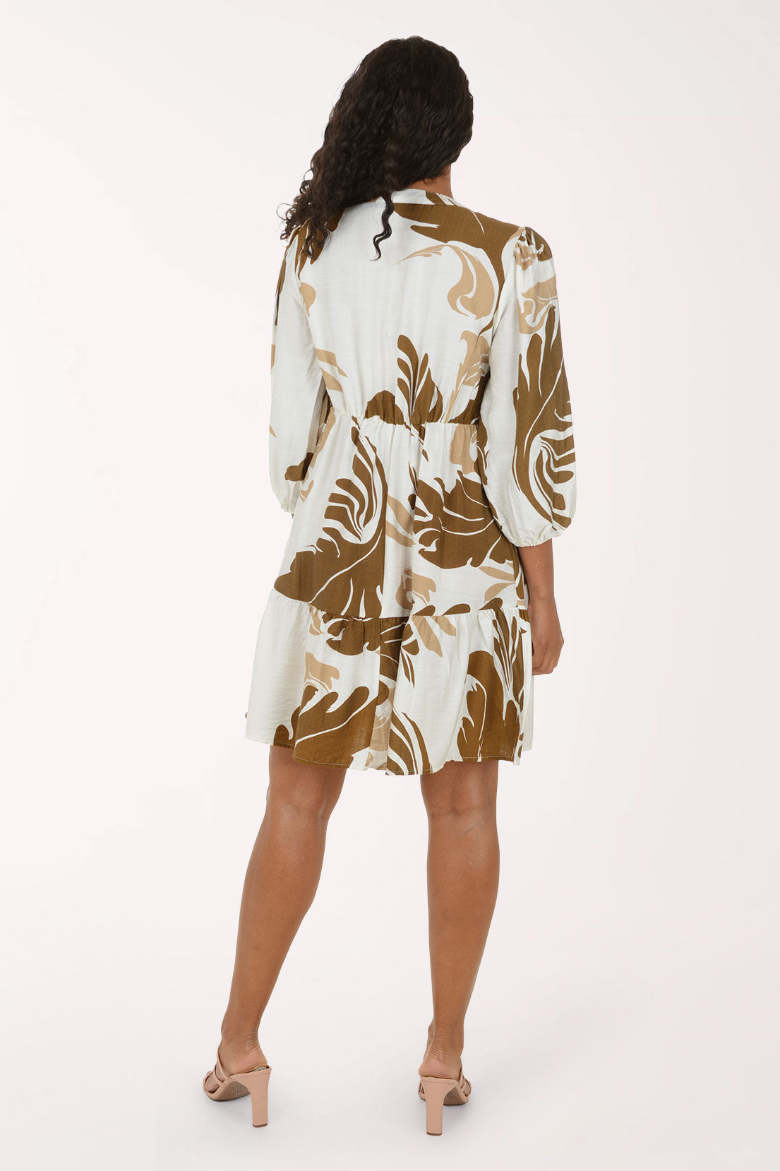 A woman modeling a short tiered dress with three-quarter sleeves, featuring an abstract botanical pattern in brown and beige on an off-white background, set against a plain white studio backdrop.
