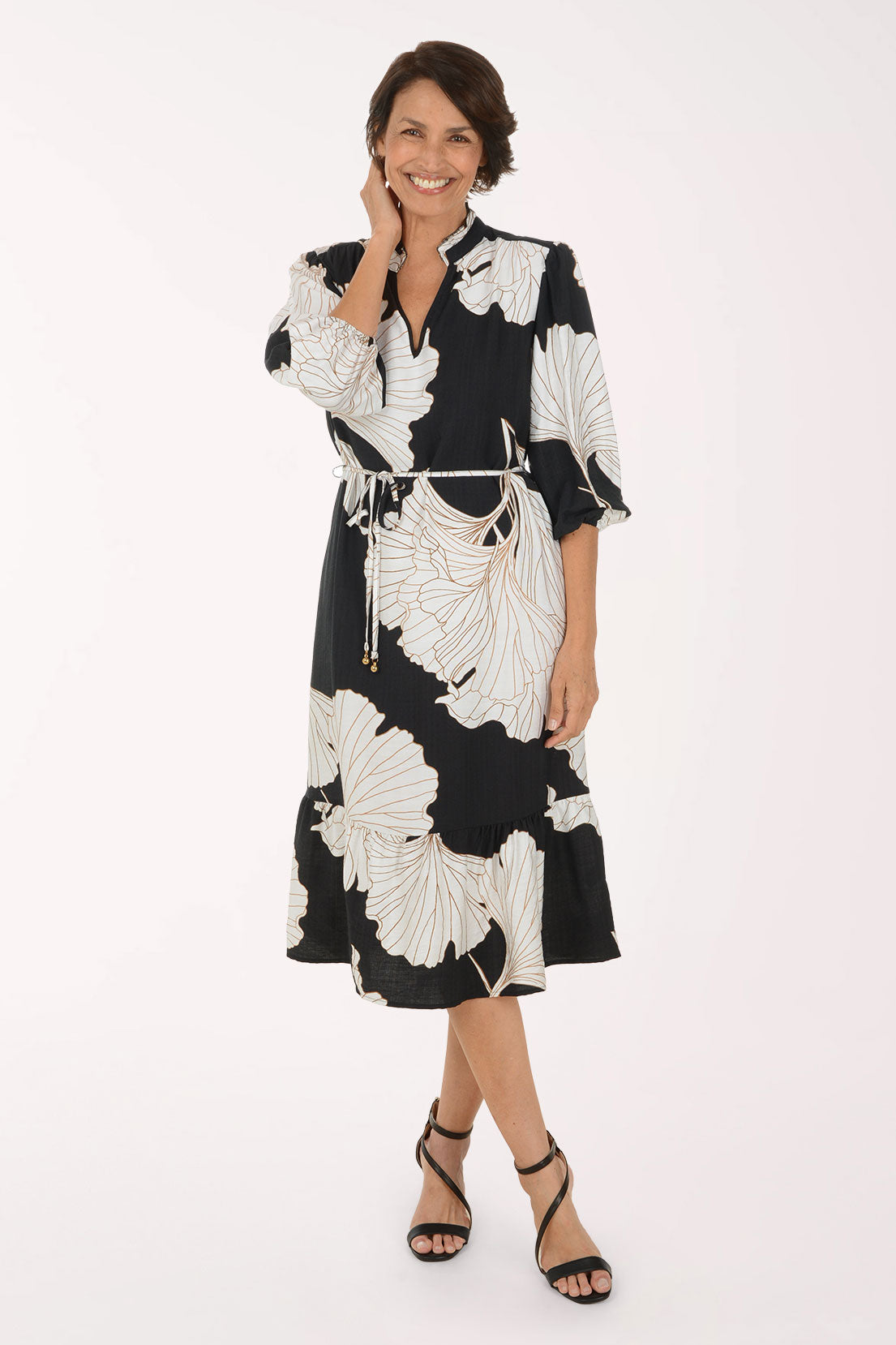 A woman modeling a black midi dress with a large white floral print, featuring a tie waist and 3/4 sleeves, set against a plain white studio background.