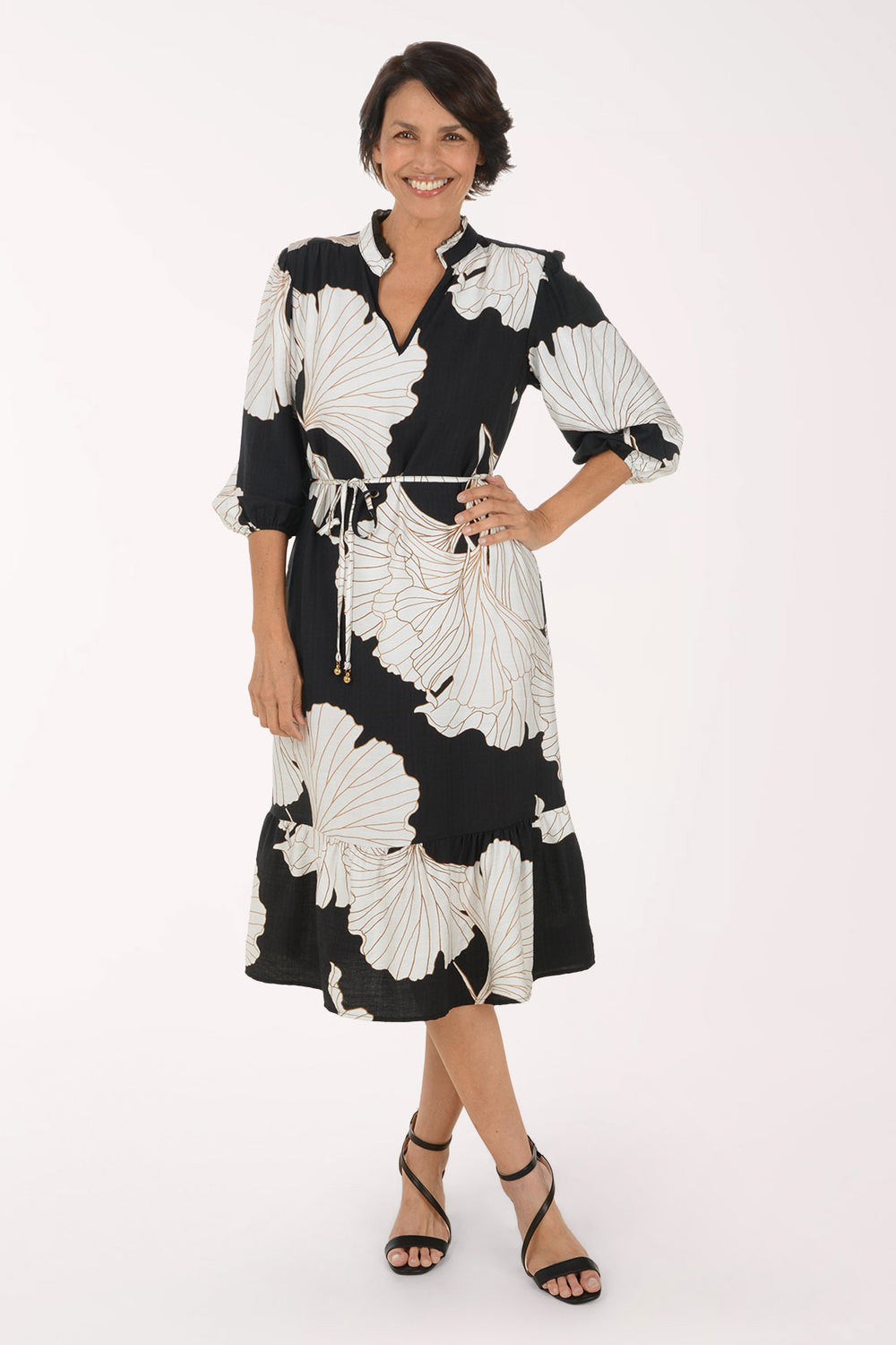 A woman modeling a black midi dress with a large white floral print, featuring a tie waist and 3/4 sleeves, set against a plain white studio background.