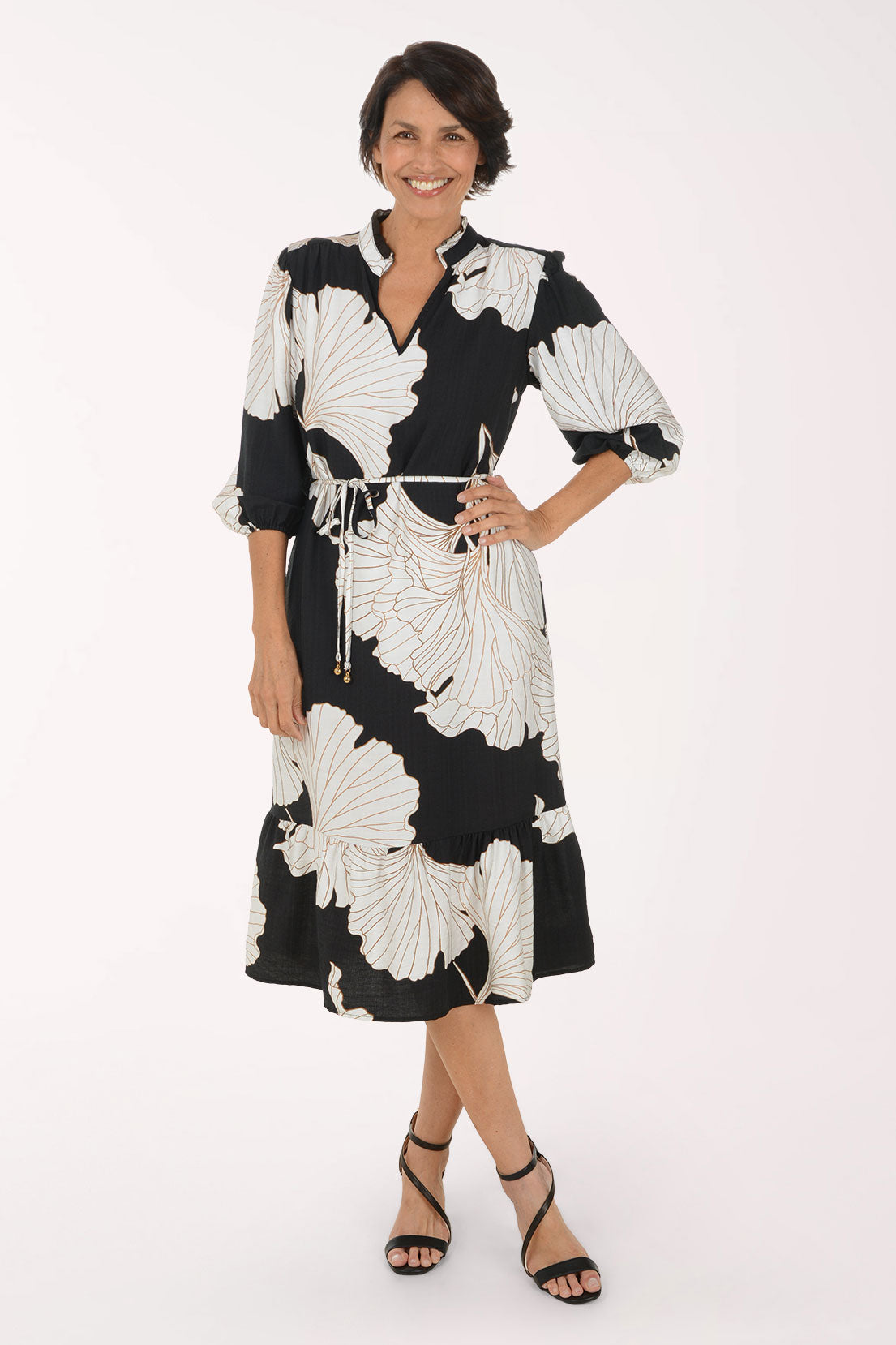 A woman modeling a black midi dress with a large white floral print, featuring a tie waist and 3/4 sleeves, set against a plain white studio background.