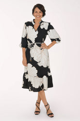 A woman modeling a black midi dress with a large white floral print, featuring a tie waist and 3/4 sleeves, set against a plain white studio background.