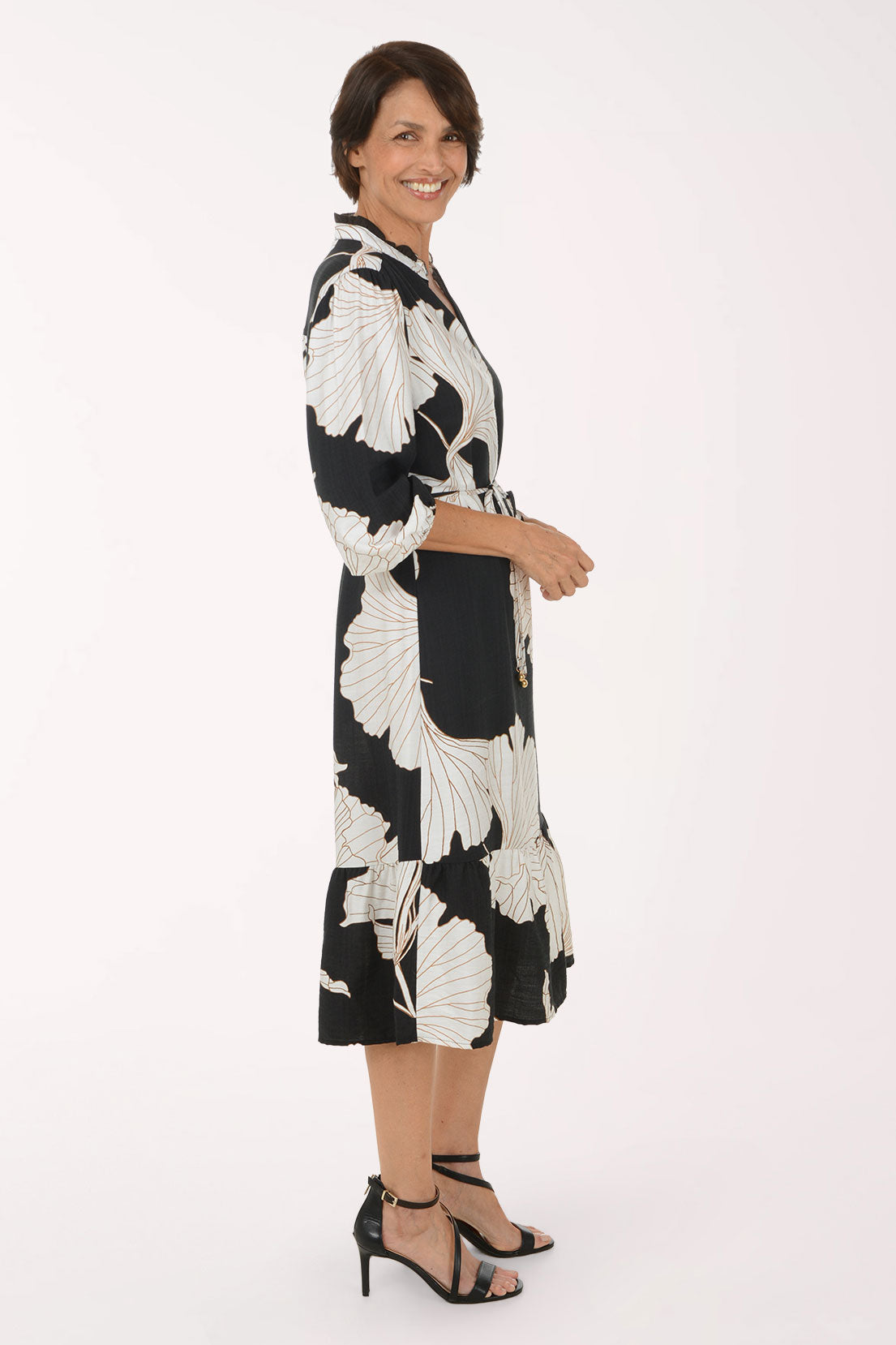 A woman modeling a black midi dress with a large white floral print, featuring a tie waist and 3/4 sleeves, set against a plain white studio background.