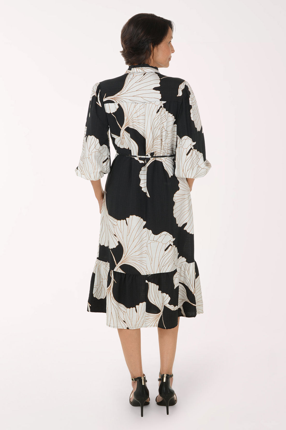 A woman modeling a black midi dress with a large white floral print, featuring a tie waist and 3/4 sleeves, set against a plain white studio background.
