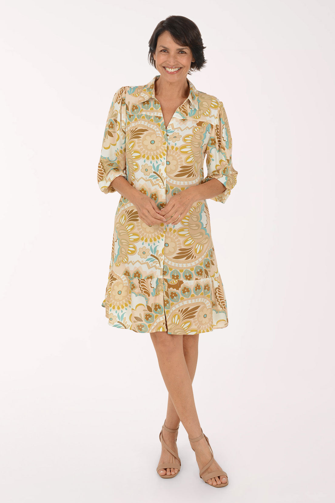 A woman modeling a short-sleeve button-up dress with a beige, gold, and light blue paisley pattern, paired with beige heeled sandals, against a plain white studio background.