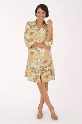 A woman modeling a short-sleeve button-up dress with a beige, gold, and light blue paisley pattern, paired with beige heeled sandals, against a plain white studio background.