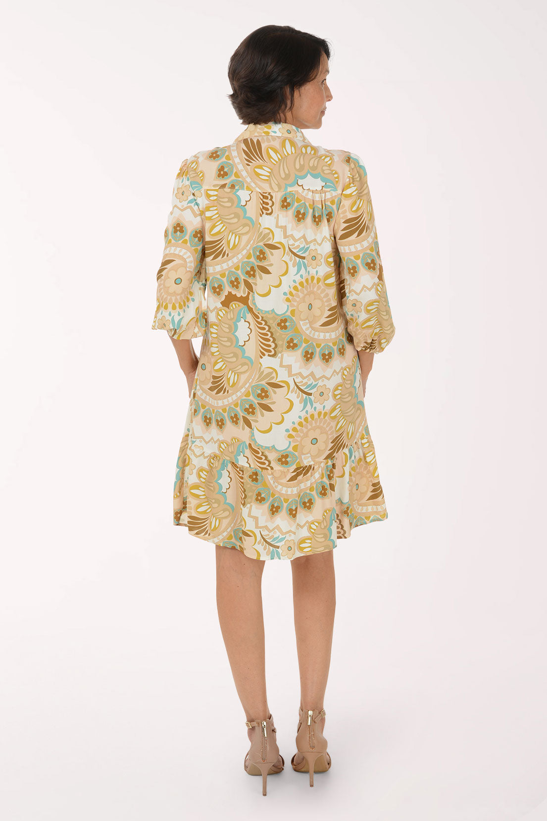 A woman modeling a short-sleeve button-up dress with a beige, gold, and light blue paisley pattern, paired with beige heeled sandals, against a plain white studio background.