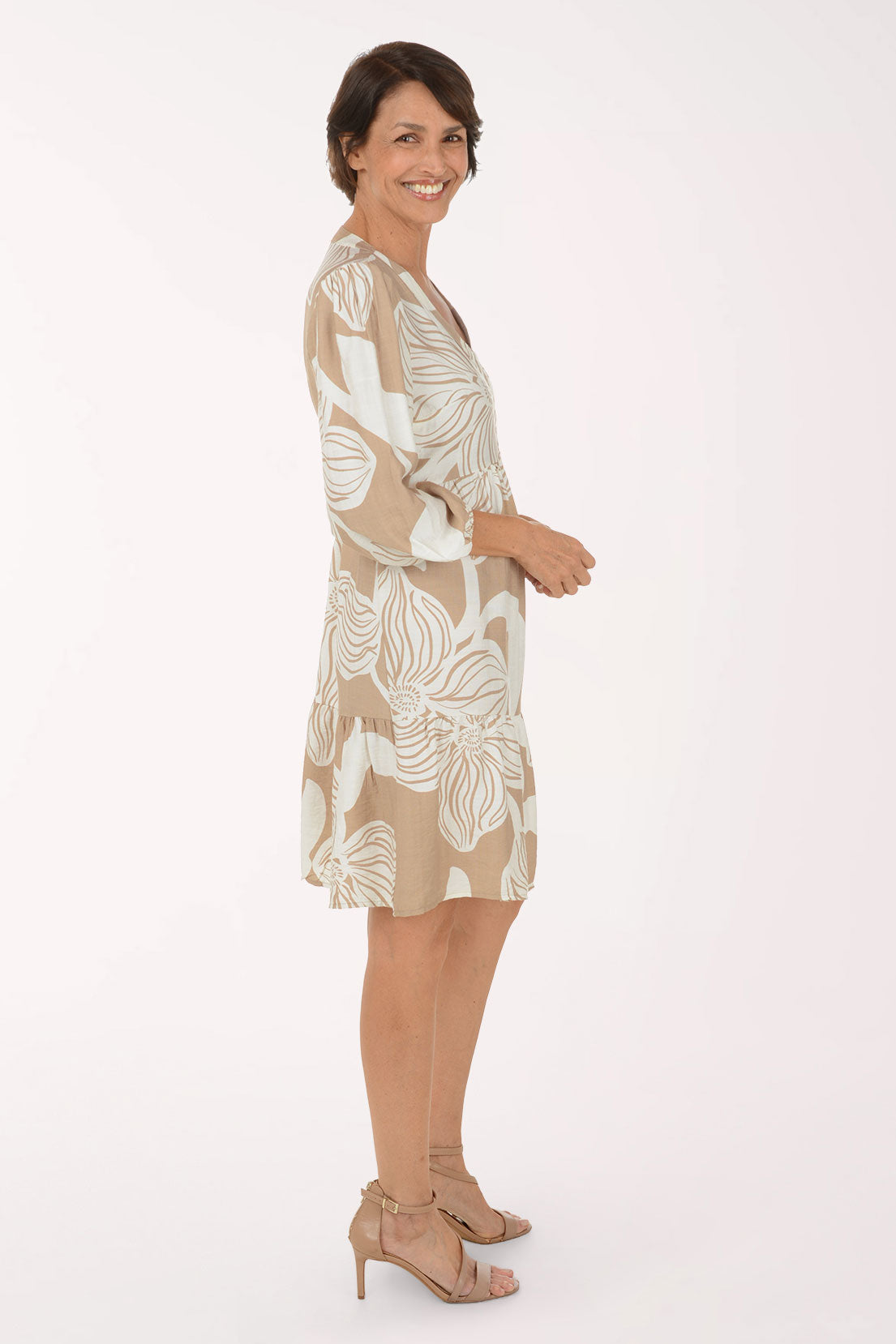 A woman modeling a beige and white tropical floral print dress with three-quarter sleeves and a V-neckline, paired with beige heeled sandals, against a plain white studio background.