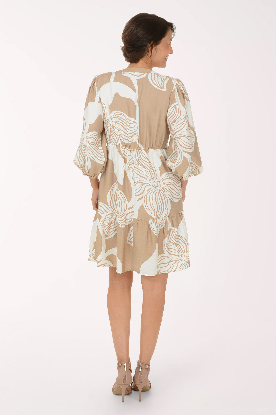 A woman modeling a beige and white tropical floral print dress with three-quarter sleeves and a V-neckline, paired with beige heeled sandals, against a plain white studio background.