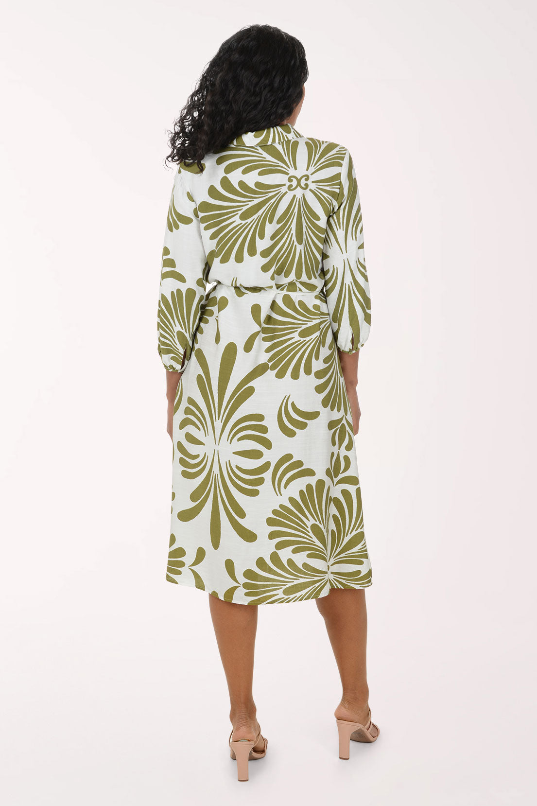 A woman modeling a white midi shirt dress with a green palm leaf pattern, featuring three-quarter sleeves, a button-up front, and a matching tie belt at the waist, paired with beige strappy heels against a solid white studio background.