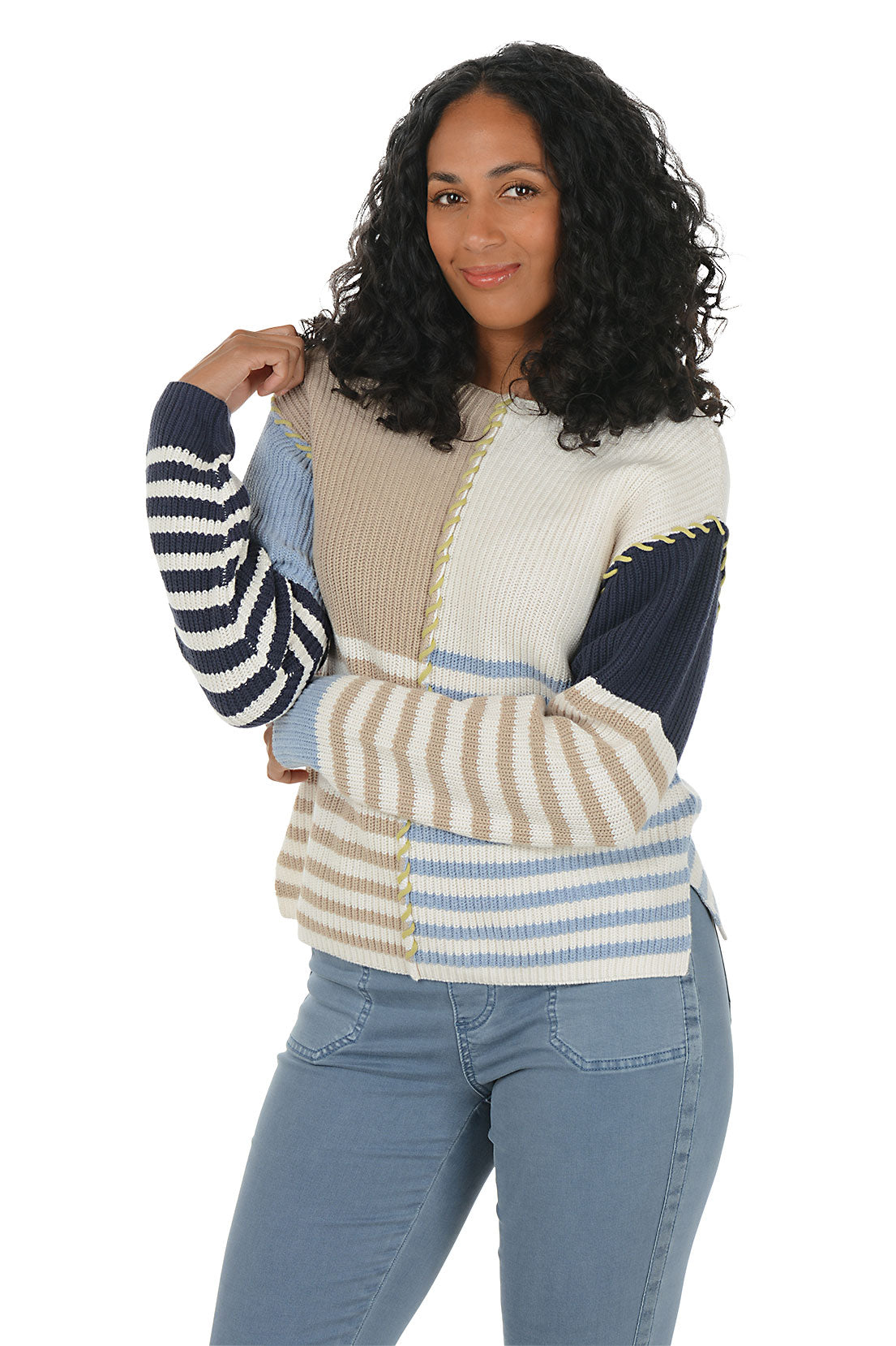 Woman wearing a colorful patchwork sweater boat neck sweater with blue jeans on a white background.