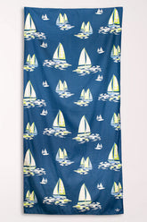 Navy blue microfiber beach towel covered in white and yellow sailboats on painted waves, shown hanging flat.