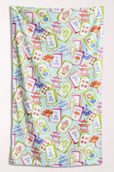 Microfiber beach towel covered in mahjong tiles, pagodas, and paper fans in pink, green, periwinkle, and coral on a pale aqua background.