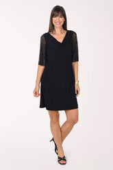 Front view of the black Tiana B Jeweled Slit Sleeve Cowl Neck Dress with a draped cowl neckline, sheer black mesh sleeves covered in small gold jewels and an above-the-knee straight hem, worn with a gold bracelet and black heels