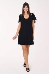 Front view of a black Tiana B Cold Shoulder Cape Dress with a scoop neckline, short cape sleeves with slits at the shoulders and an above-the-knee straight hem, styled with a gold cuff bracelet and black strappy sandals