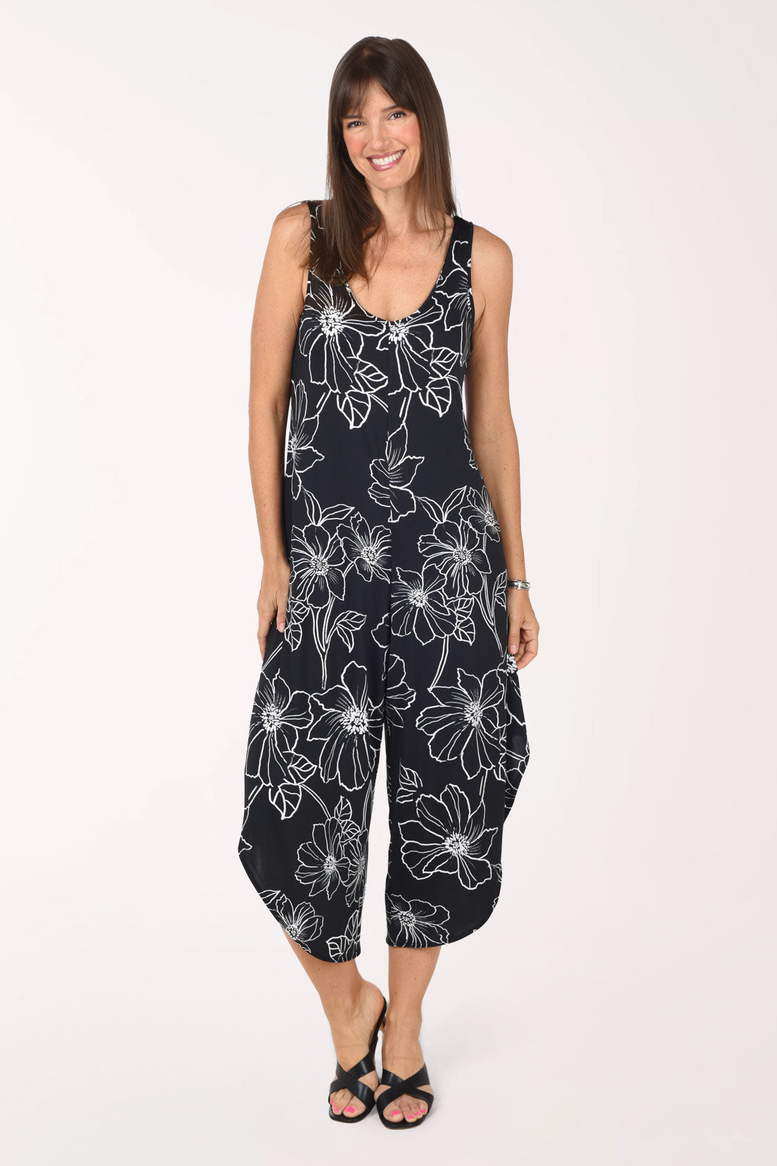 Front view of the Tiana B Black Floral Sleeveless Wide Leg Jumpsuit, black with large white outlined flowers, sleeveless with a rounded neckline and loose wide legs ending in curved, angled hems just below the knee, worn with black slide sandals