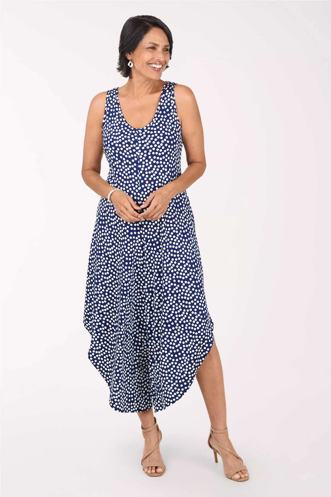 Front view of the Tiana B Navy Polka Dots Sleeveless Wide Leg Jumpsuit, navy with scattered white dots, a V-neckline and wide legs that fall like a skirt with curved, angled hems below the knee, worn with nude heeled sandals