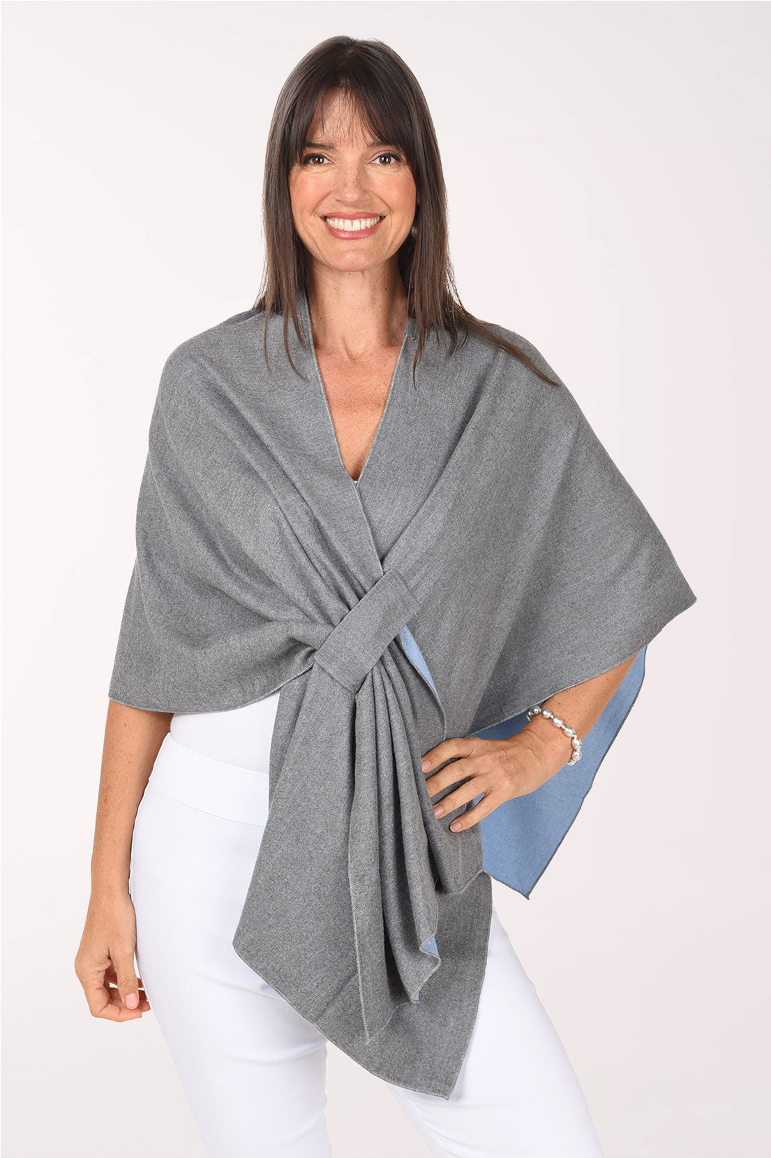 Front view of a woman in a heather grey knit wrap pulled through its front keyhole loop, showing a sky-blue contrast lining at the edges, worn over a white top with white pants.