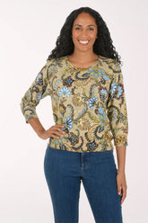 A smiling woman with long curly black hair models a three-quarter sleeve top with a detailed paisley and floral pattern in blue, green, gold, and brown on a beige background, paired with blue jeans against a plain white studio background.
