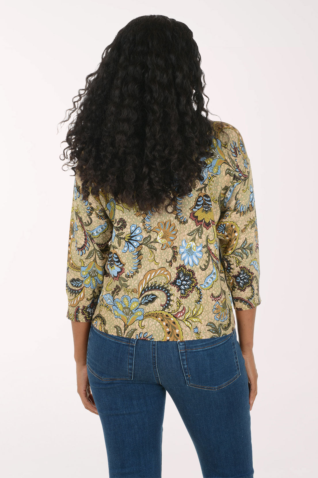 A smiling woman with long curly black hair models a three-quarter sleeve top with a detailed paisley and floral pattern in blue, green, gold, and brown on a beige background, paired with blue jeans against a plain white studio background.