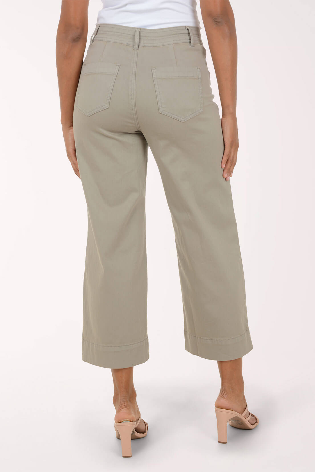 A model wearing a pair of light sage green cropped pants with a rolled hem, paired with beige strappy high-heeled sandals, against a plain off-white studio background.