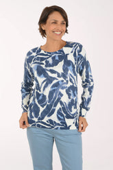 A woman modeling a long-sleeve top with a blue and white abstract pattern, paired with light blue pants, against a plain white studio background.