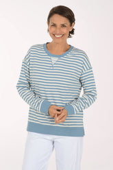 A smiling woman wearing a light blue and white striped long-sleeve henley top with white pants, standing against a plain white studio background.
