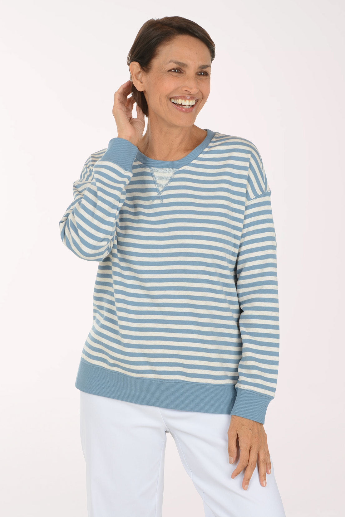 A smiling woman wearing a light blue and white striped long-sleeve henley top with white pants, standing against a plain white studio background.