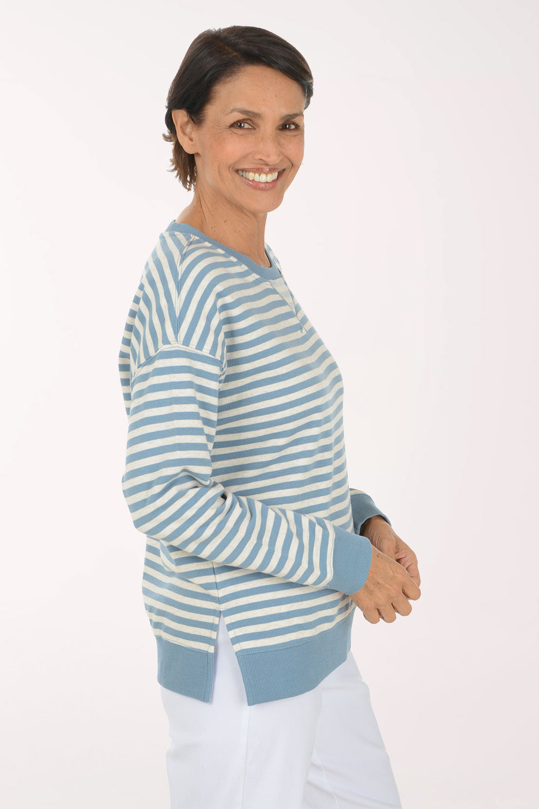 A smiling woman wearing a light blue and white striped long-sleeve henley top with white pants, standing against a plain white studio background.