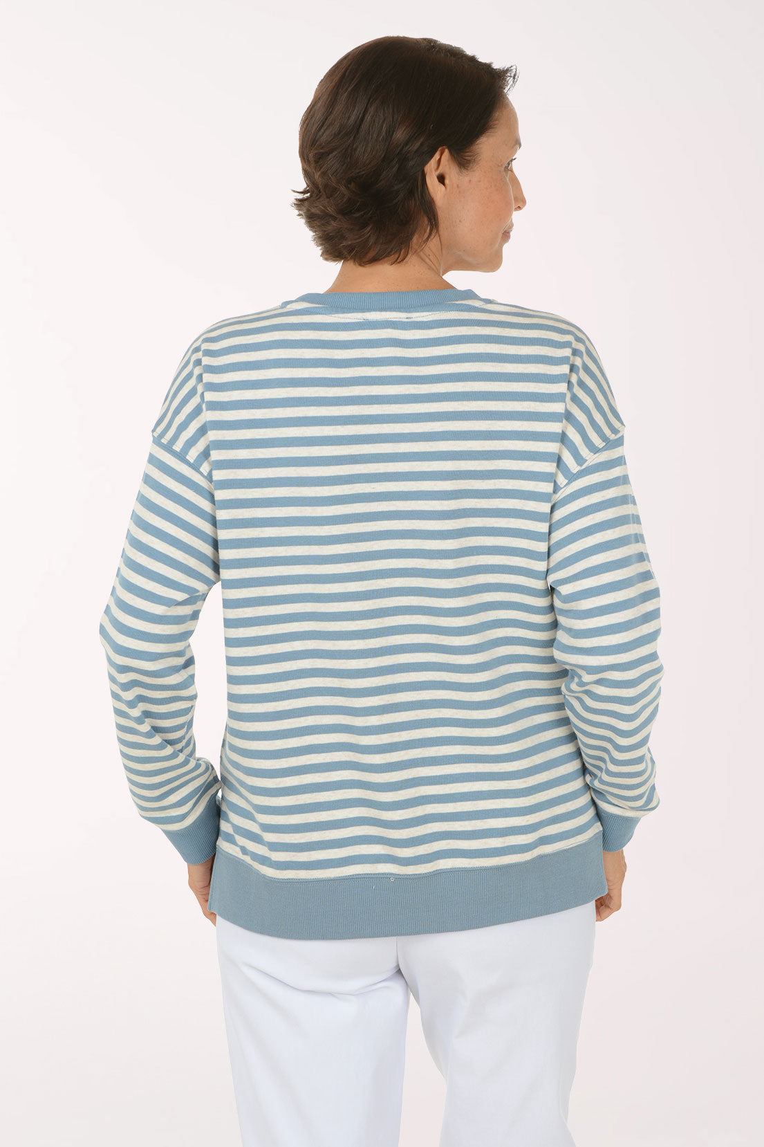 A smiling woman wearing a light blue and white striped long-sleeve henley top with white pants, standing against a plain white studio background.